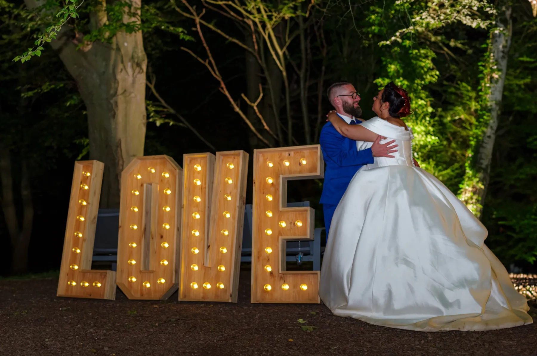 De nuit, les mariés enlacés à côté de grandes lettres lumineuses « LOVE »