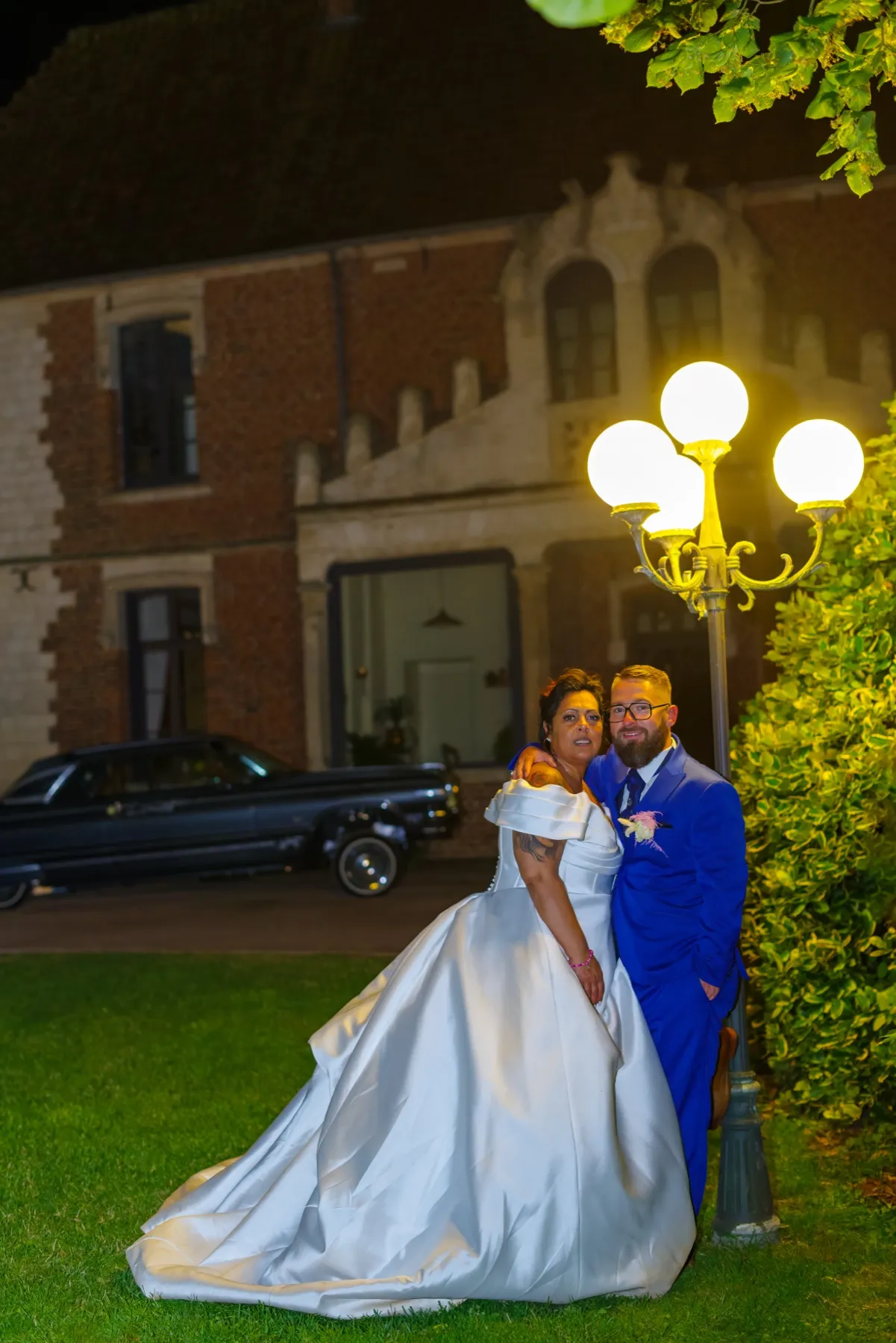 De nuit, les mariés posent sur la pelouse sous un lampadaire, une voiture ancienne derrière eux
