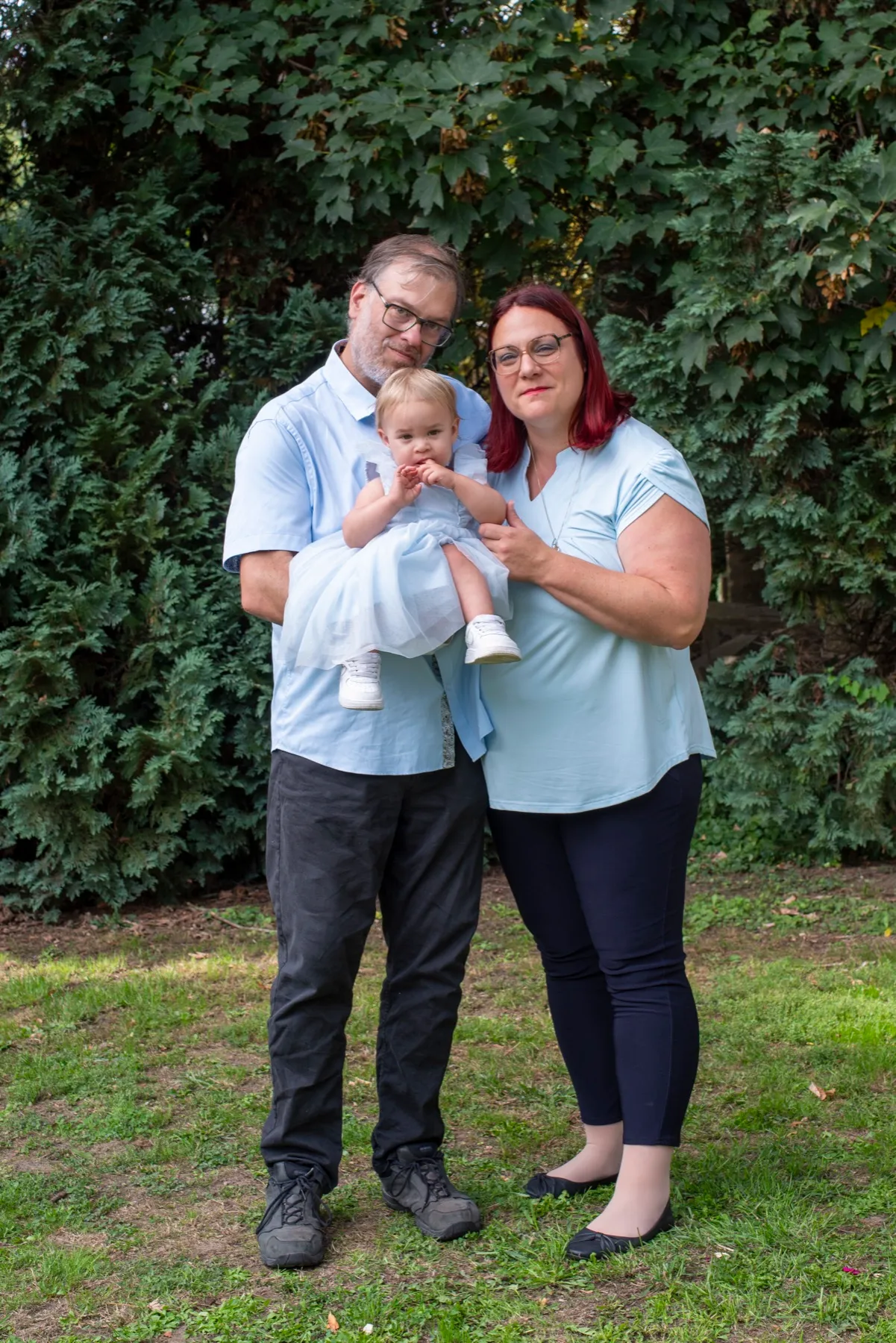 Un couple pose avec son bébé dans les bras, devant la haie du jardin