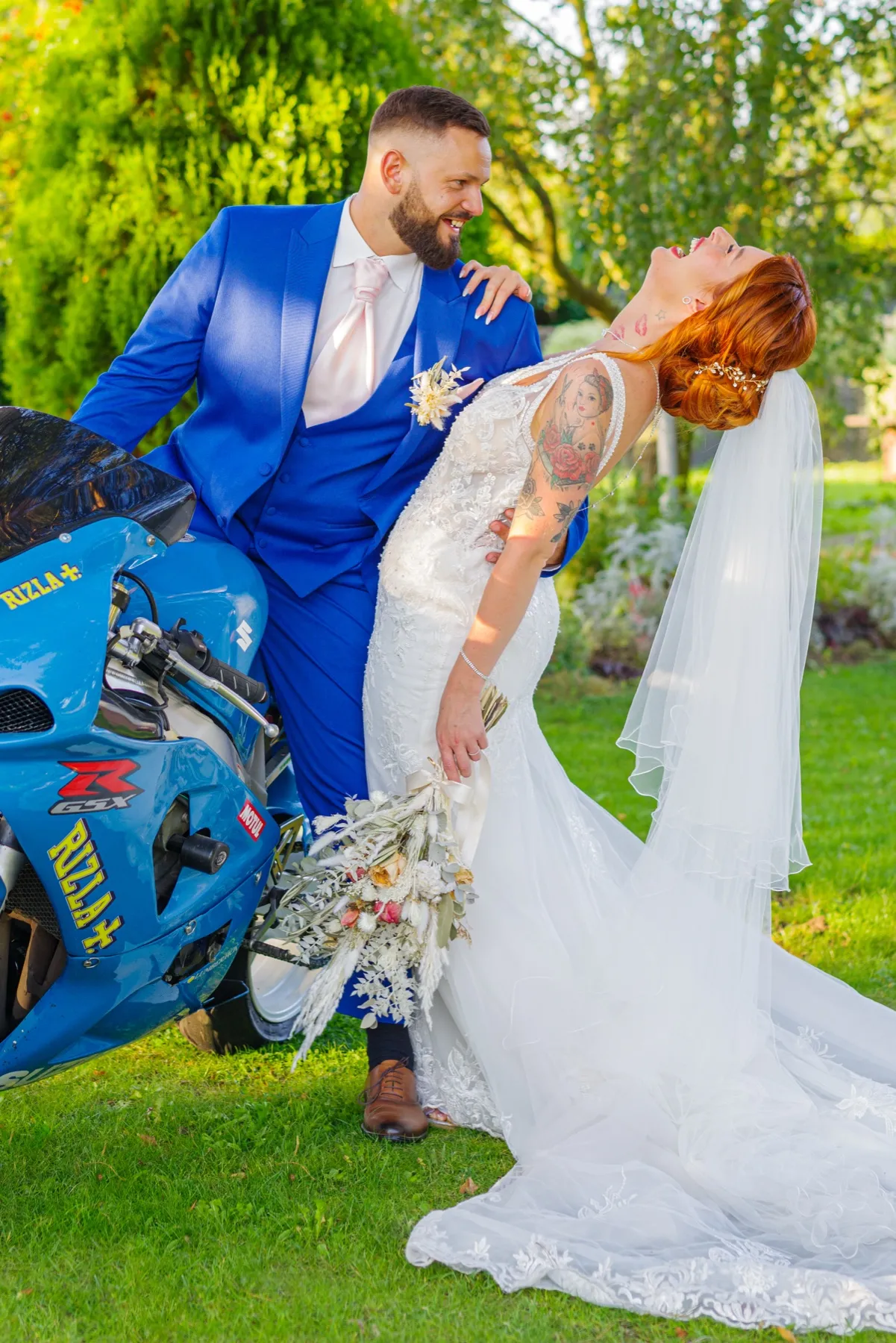 La mariée, renversée en arrière, éclate de rire dans les bras du marié en costume bleu, près d'une moto